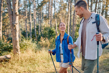 Young couple walking through a vibrant forest, using trekking poles and soaking up the sun on a...