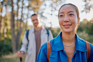 Young Asian woman smiling while hiking along a scenic forest trail, with a blurred figure of a man enjoying the outdoor adventure in the background