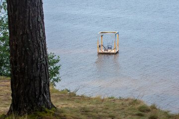 A serene wooden dock extends into a calm lake, featuring cozy swing and seating area under simple canopy, surrounded by lush greenery and trees