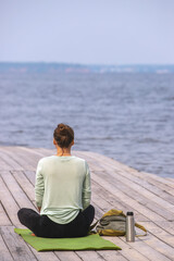 A woman sits in a meditative pose on yoga mat by the water, enjoying peaceful moment of mindfulness and relaxation under serene sky