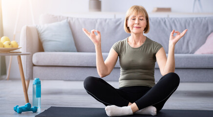 Full length of mature woman meditating with closed eyes, making gyan mudra with her hands at home....