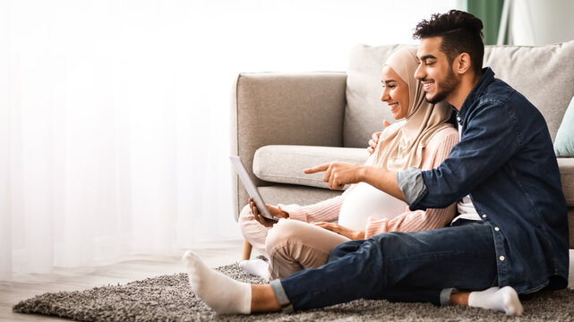 Happy islamic family man and pregnant woman using laptop together at home, cheerful middle eastern spouses making online shopping for future baby while relaxing with computer on floor in living room