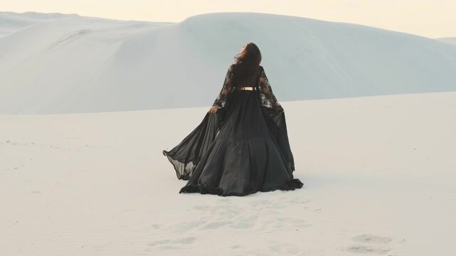 silhouette beautiful woman in silk black dress with long train. fabric brunette dark hair flies in wind blow slow motion. fantasy girl walks in desert white sand. sunset sky sun light back rear view.