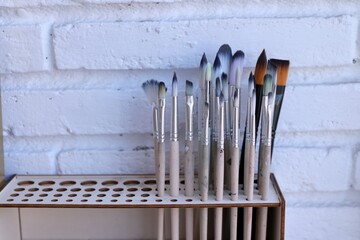 Assortment of Clean Artist Paintbrushes in Wooden Holder Against White Brick Wall