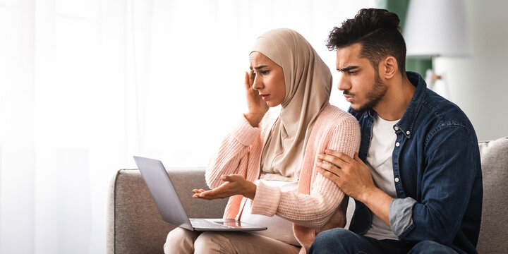 Labor And Delivery Fears. Worry Muslim Couple Watching Childbirth Videos On Laptop While Sitting Together On Couch At Home, Concerned Muslim Woman In Hijab Having Prenatal Anxiety, Copy Space - Powered by Adobe