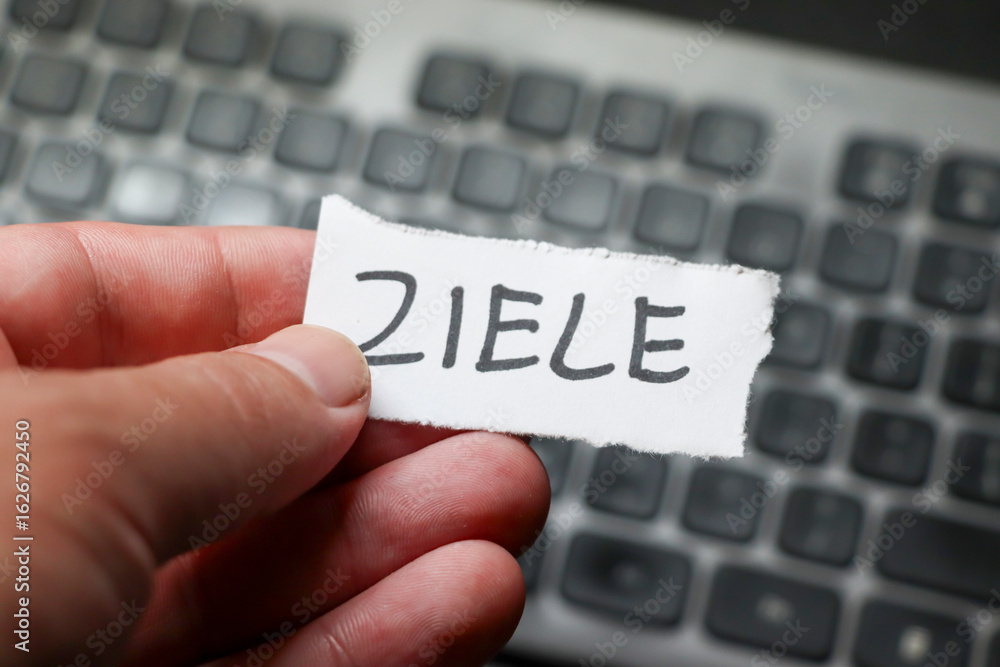 Poster a person holding a piece of paper with the word ziele while sitting in front of a computer keyboard,