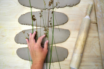 A hand arranges delicate plants onto rolled clay slabs, preparing for botanical imprints. This step...