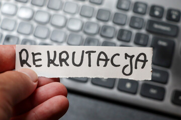 Naklejka premium A hand holding a sign reading REKRUTACJA on a blurred computer keyboard background, symbolizing recruitment processes in modern workplaces.