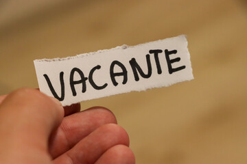 A person holding a torn piece of paper with the word VACANTE written in bold letters. The background is blurry, emphasizing the message, possibly indicating a vacancy sign in a business or rental con