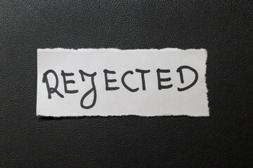 A torn paper note with the word REJECTED written in bold, creative font, placed against a dark,...