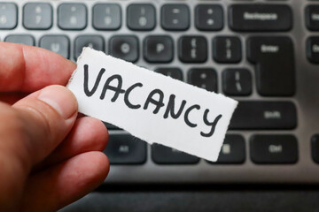 A hand holding a sign reading VACANCY in front of a laptop keyboard, indicating job opportunities...