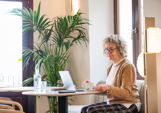 Middle-aged woman working remotely with coffee in hand at cozy café, taking a mindful pause while seated by the window with laptop in bright and peaceful atmosphere