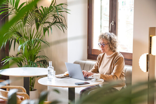 Middle-aged woman working remotely on laptop from cozy café during vacation, enjoying peaceful atmosphere and natural light while balancing work and leisure
