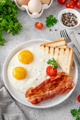 Breakfast with fried eggs, bacon and toasts on a white plate with cup of coffee on a stone background. Top view, selective focus.