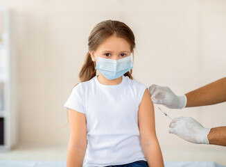 Little girl in face mask being vacciniated against covid-19 at hospital. Cute kid getting...