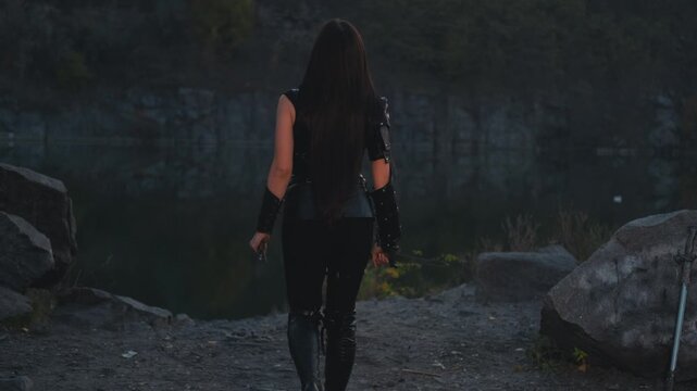 silhouette fantasy woman warrior assassin holds dagger in hands walks at night nature. Rear view, back armed girl. Black leather costume, hood. Ninja soldier with sword knife. 4k footage. river rocks.