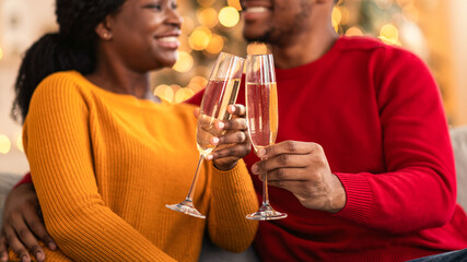 Relationship, love, celebrating New Year at home together during lockdown. Cheerful millennial african american man and woman clink glasses of champagne in interior with Christmas tree with garlands