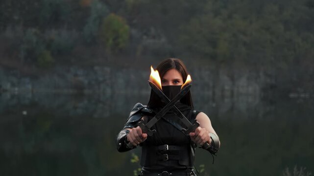 Portrait fantasy woman warrior assassin holding two burning blade daggers in hands with bright flame fire. hiding face behind mask. Armed warlike girl. Black leather costume ninja soldier. 4k footage