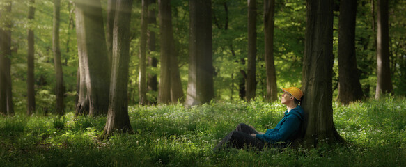 Young man sitting under trees in the forest and looking into the gentle tranquil rays of sunlight. Panoramic format.