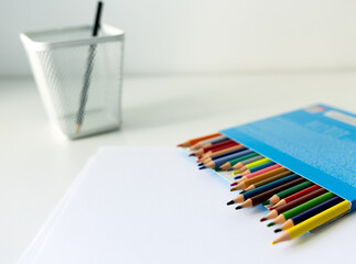Fototapeta premium schoolboy workplace: white desk with stand with colored pencils and sketchbook. school supplies on the table beautifully illuminated by a desk lamp: colored pencils in a stand and a sketchbook