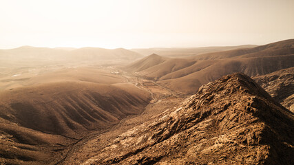 Aerial view of a vast, dry mountain landscape on Fuerteventura bathed in warm evening light. The...