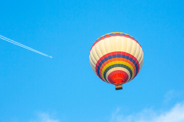 Colorful hot air balloon ascends in clear blue sky near airplane leaving vapor trail