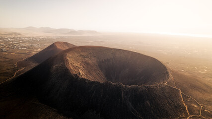 Aerial drone shot of a dramatic volcanic crater at sunset on Fuerteventura. The warm golden light highlights the textures and tones of the barren landscape, creating a cinematic and atmospheric scene.