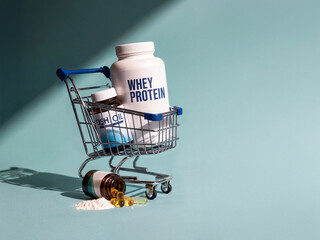 Whey protein and fish oil supplements in mini cart with capsules and powder spilled on pastel surface under angled lighting