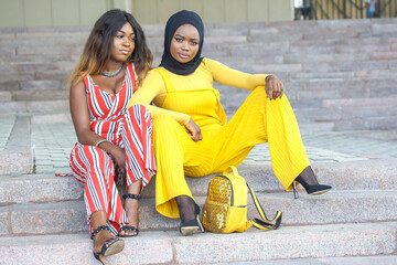 Two African women in stylish outfits pose together on outdoor steps in bright, vibrant colors