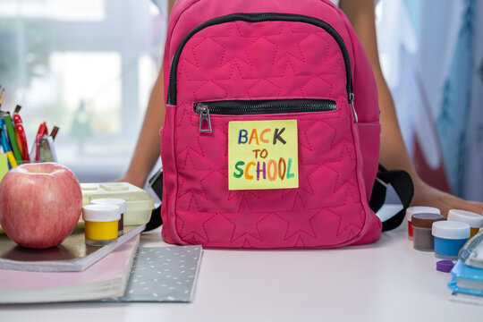 On a table, a pink school backpack with a back to school note stands among various stationery items.  - Powered by Adobe