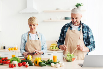 Adult couple prepare food together for family dinner. Smiling mature man and woman make salad in kitchen interior with bright vegetables, glasses of wine and laptop on table, empty space, panorama
