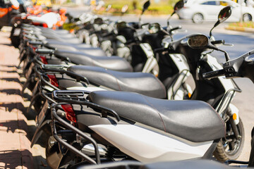 Scooters line a sidewalk in a bustling city, catching sunlight as cars drive past. The scene...