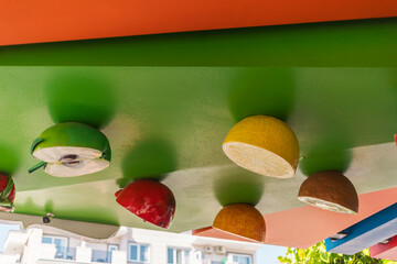 Brightly colored cut-out fruits dangle from a green awning at an outdoor market. The atmosphere is...