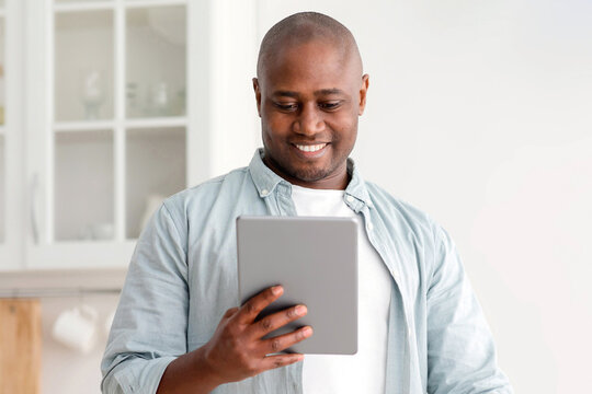 Positive african american mature man reading news on digital tablet and drinking coffee in kitchen, browsing social networks while enjoying hot drink at home, free space