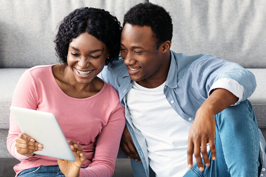 Smiling black couple with cleaning tools resting, sitting on floor in living room, using digital tablet, looking for house-keeping service on Internet. Tired african american man and woman with pad
