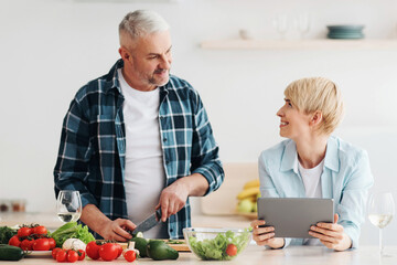 Cooking in kitchen together, searching recipe in internet, watch online blog. Happy middle aged man cuts vegetables for salad, woman with tablet look food lesson in kitchen interior, empty space