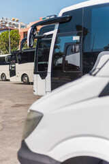 A variety of buses in different colors are parked at a travel terminal on a sunny day. The vehicles...
