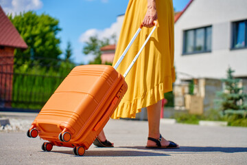 Woman in yellow dress stands outdoors holding orange suitcase near residential house in suburban...
