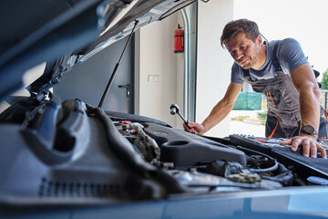 Male mechanic working on car engine with wrench under open hood in garage. Man in work overalls is...