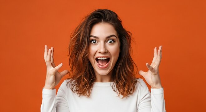 Young woman expresses extreme joy and excitement with wide eyes and open mouth against a vibrant orange background