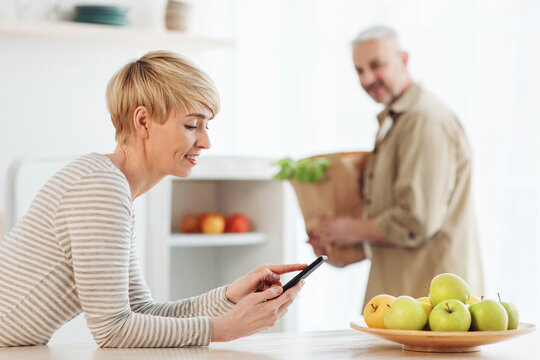 Payment for online delivery of products, review and rating of service. Happy senior man unpacks package with vegetables, lady typing at smartphone on modern light comfort kitchen interior, free space