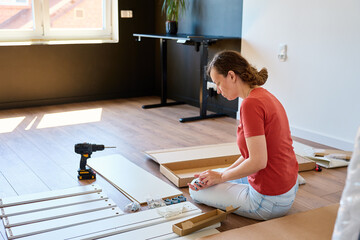 Woman unpacking parts for furniture assembly at home. Female sitting on floor and holding bed...