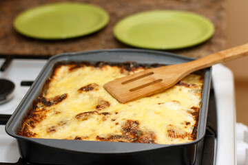 Baked lasagna in a black tray on a stove. A wooden spatula lies on top