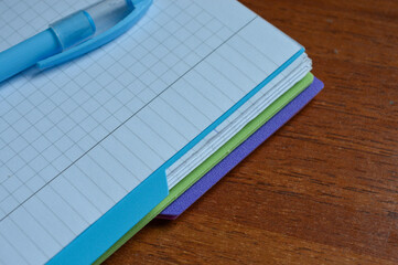 a stack of squared notebooks with a blue pen lying on a wooden desk, top notebook is spiral-bound...