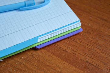 a stack of squared notebooks with a blue pen lying on a wooden desk, top notebook is spiral-bound...