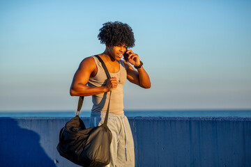 Young mixed race man on a phone call holding gym bag by the seaside