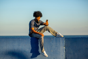 Smiling African American young man taking a selfie while sitting on blue wall by the sea