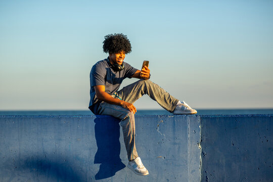 Smiling African American young man taking a selfie while sitting on blue wall by the sea