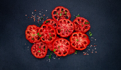 Freshly sliced ripe tomatoes arranged on a textured dark surface with herbs and salt scattered...