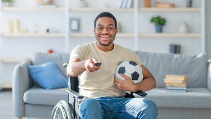 Cheerful black guy in wheelchair watching football game on TV at home. Sports fan with disability...